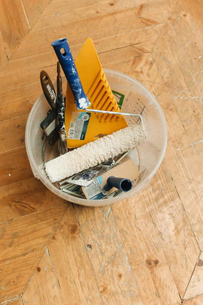 Top view of various painting tools in a bucket on a wooden floor.